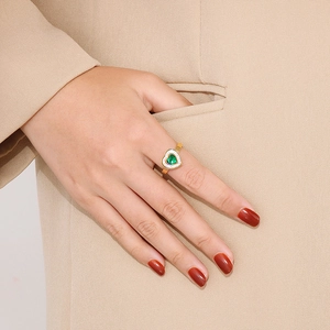 Detail of 18k gold heart ring with green stone worn on finger against a beige blazer