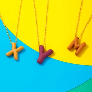 Minimalist gold necklace with matte textured X, Y, and M letter pendants on a colorful background