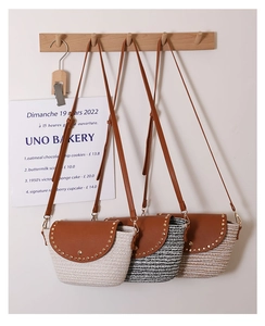 Detail of crossbody straw bags with PU leather flap and rivet accents on a wooden hook