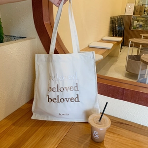 Gros plan du sac fourre-tout beige en toile avec broderie beloved sur table en bois