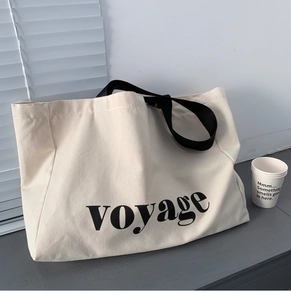 Detail of off-white canvas tote bag featuring black voyage lettering and a wide strap