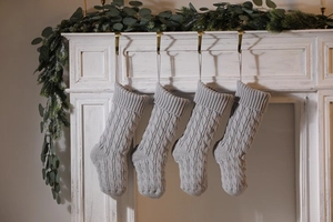 Detail of four gray cable-knit Christmas stockings hanging on a white mantel with greenery