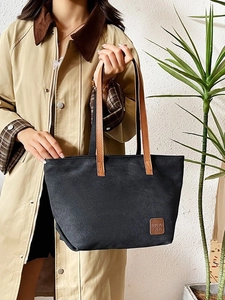 Minimalist black canvas tote bag with brown leather straps shown on a beige trench coat