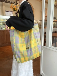 Large yellow and purple plaid canvas tote bag worn by a model in a cozy cafe setting