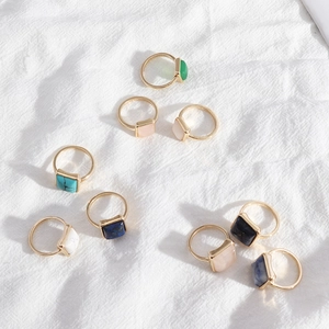 Flat lay of colorful square stone rings in gold finish on white linen fabric texture