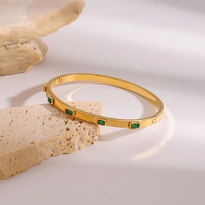 Gold plated stainless steel bangle with five emerald-cut green zircons on stone background