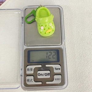 Miniature green garden shoe keychain being weighed on a digital scale at 12.2g