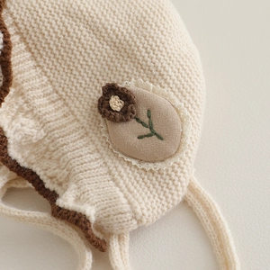 Off-white knit baby bonnet with brown scalloped edges and floral appliqué detail