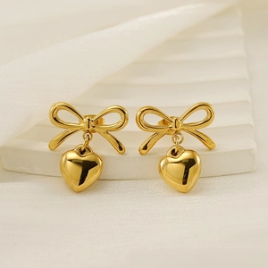 Detail of gold bow and heart drop earrings on a white stand with soft fabric background