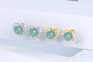 Detailed view of silver and gold heart earrings with green gemstones on white pedestals
