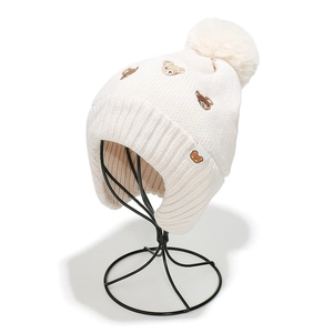 Détail du bonnet en tricot blanc cassé avec broderie ours, cache-oreilles et pompon en fourrure