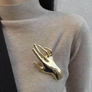 Detailed view of a gold metallic hand-shaped brooch pinned to a beige ribbed sweater