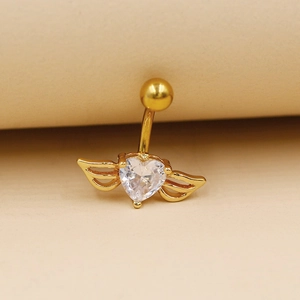 Détail macro d'un piercing nombril doré avec cœur et ailes en zircone sur fond beige
