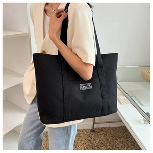 Minimalist black canvas tote bag shown on shoulder of a model in white blazer