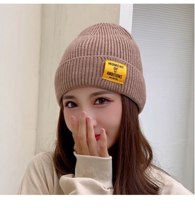 Close-up of a brown knit beanie with a yellow logo patch worn by a model in bright daylight