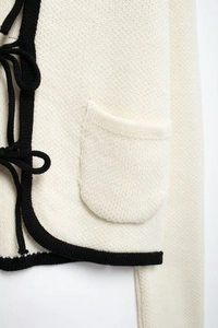 Détail du cardigan en maille crème avec bordure noire contrastée et nœud élégant
