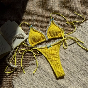 Flat lay of yellow crochet bikini with white lace trim and light blue bows on woven mat