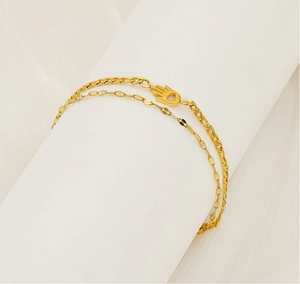 Detail of golden double-layer chain bracelet featuring a Hamsa hand charm on a white stand