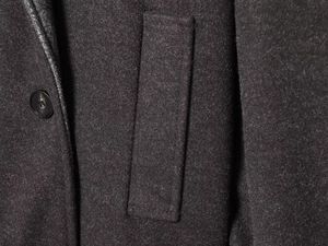 Detail view of a black round button and a straight pocket on dark grey wool coat fabric
