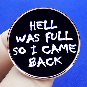 Detail of round black enamel pin with white text HELL WAS FULL SO I CAME BACK in rose gold frame