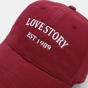 Close-up of LOVE STORY EST 1989 white embroidery on a burgundy cotton baseball cap