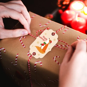 Detail of hand tying a cartoon fox Christmas gift tag on a kraft paper box