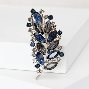 Detail of silver alloy leaf brooch with blue and grey crystals on a white pedestal