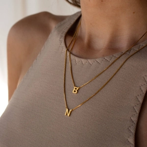 Gold box chain necklaces with letter B and N pendants worn by a model in soft lighting