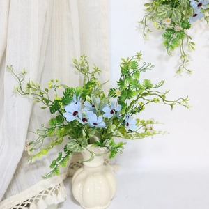 Artificial blue daisy flowers in a white ceramic vase with green leaves home decor