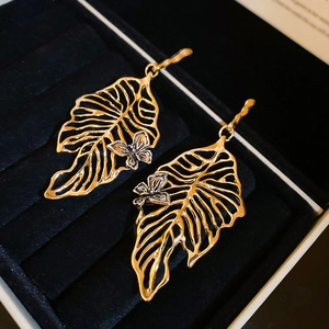 Close-up of golden monstera leaf earrings with silver butterfly detail on black velvet