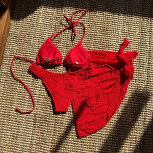 Flat lay of red three-piece bikini set with textured fabric on a woven mat background