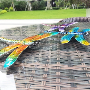 Three colorful metal dragonfly wall art pieces displayed on a modern outdoor glass patio table