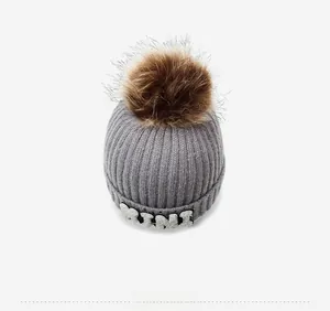 Top view of a grey knit beanie with a brown faux fur pompom and MINI letter patch