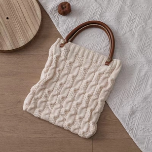 Cream knit tote bag with cable patterns and wooden handles on a lace surface