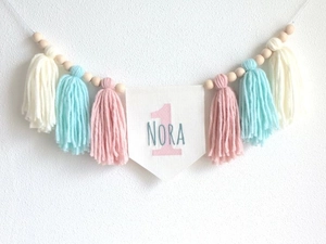 Detail of handmade first birthday banner with pink fringe, wood beads, and name Nora