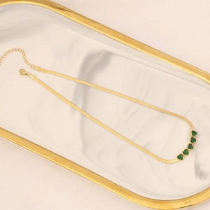 Gold snake chain necklace with five emerald green heart gems on marble tray
