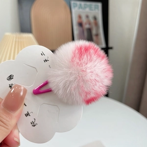 Close-up of a mini plush hair clip with white and pink pom poms on a floral card