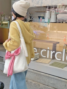 Lifestyle shot of a white plush tote bag with a red plaid bow on a yellow hoodie model