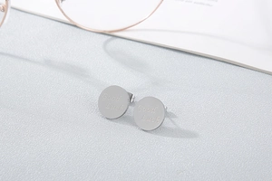 Close-up of silver steel stud earrings featuring engraved Good Luck letters on textured background