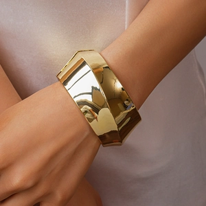 Detailed close-up of a wide geometric faceted gold-finished metal bangle on a model wrist
