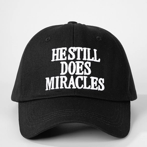 Close-up of white 3D embroidery text He Still Does Miracles on a black cotton baseball cap