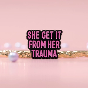 Macro detail of enamel brooch with pink SHE GET IT FROM HER TRAUMA text and pearls