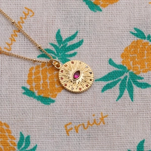 Detail of gold evil eye necklace with sunburst texture and red zircon on summer fabric