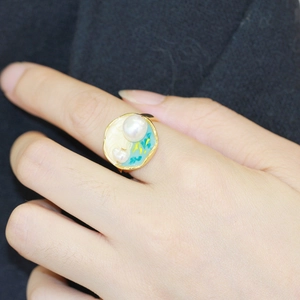 Gros plan d'une bague dorée avec perle et motif océan en émail bleu au doigt
