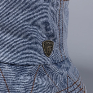 Detail of light blue denim bucket hat featuring vintage metal badge and orange stitching