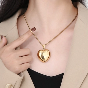 Close-up of a large hollow heart pendant on a gold snake chain worn over a beige blazer
