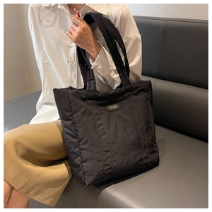 Detail view of black quilted nylon tote bag with metal logo on a soft grey sofa texture