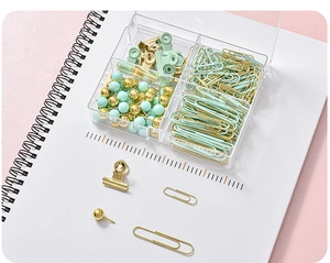 Close-up of mint green and gold binder clips, paper clips, and push pins in a clear box