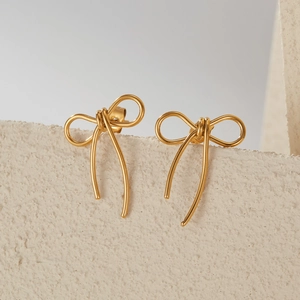 Minimalist gold bow stud earrings in stainless steel on a textured neutral background