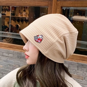 Beige knitted beanie hat featuring a cute cartoon cat heart badge on a female model